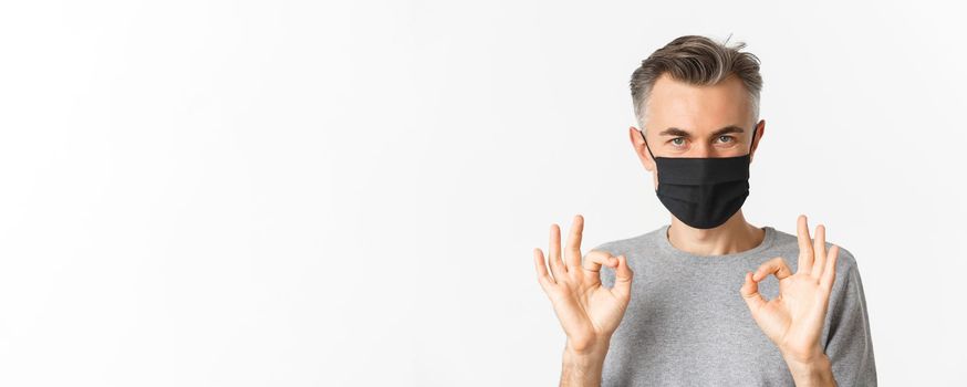 Concept of covid-19, social distancing and quarantine. Close-up of confident middle-aged man, wearing black medical mask, assure everything good, showing okay signs, white background.