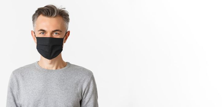 Concept of covid-19, social distancing and lifestyle. Close-up of handsome middle-aged man in black medical mask, looking confident at camera, standing over white background.