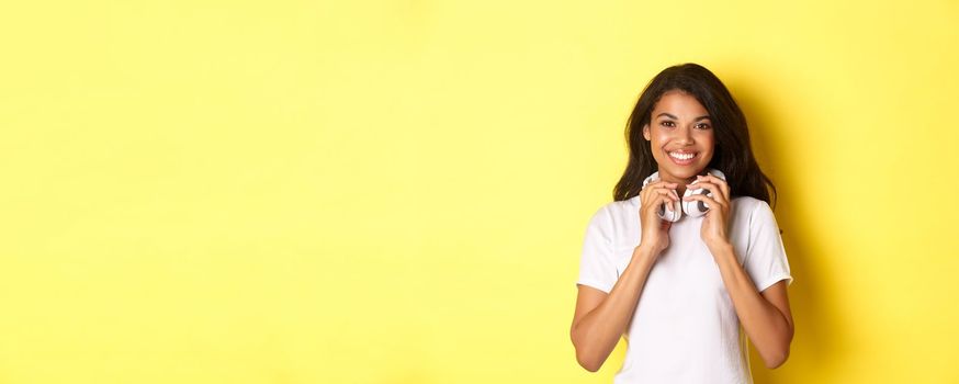 Portrait of beautiful teenage african-american girl, holding headphones on neck and smiling at camera, standing over yellow background.