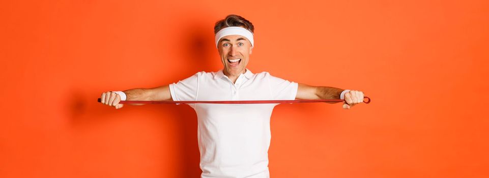 Concept of workout, gym and lifestyle. Active and healthy middle-aged fitness guy, holding stretching rope for exercises and smiling, standing over orange background.