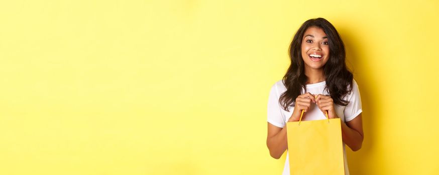 Portrait of cheerful african-american girl shopping, opening a bag with gift and smiling happy, standing over yellow background.