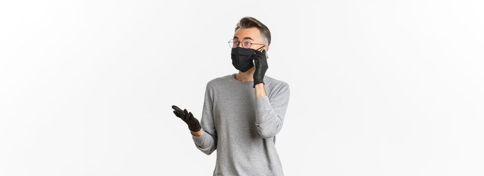 Concept of covid-19, social distancing and lifestyle. Image of handsome middle-aged man in medical mask and gloves, talking on mobile phone, standing over white background.