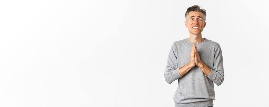 Image of nervous and desperate middle-aged man begging for help, holding hands in pray and looking worried, need favour, standing over white background.