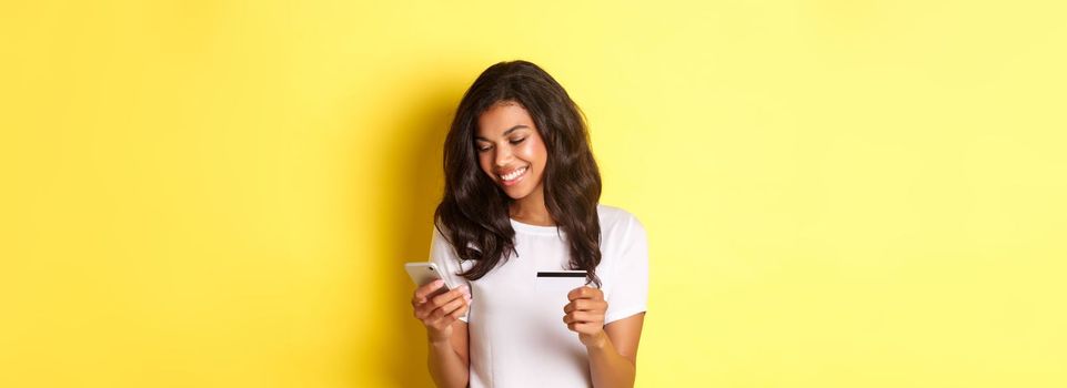Portrait of good-looking african american woman, shopping online with smartphone and credit card, standing over yellow background.