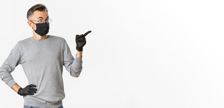 Concept of coronavirus, lifestyle and quarantine. Portrait of surprised handsome middle-aged man looking and pointing right, wearing black medical mask and gloves, standing over white background.