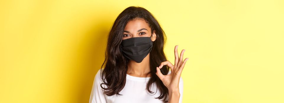Concept of coronavirus, pandemic and lifestyle. Close-up of pretty african-american woman in black face mask, showing okay sign in approval, praise good work, yellow background.