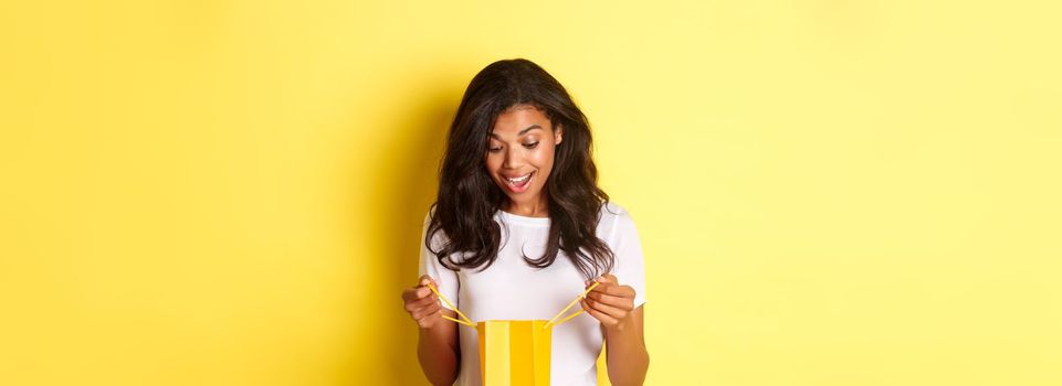 Image of happy and surprised african-american girl, receiving a gift, looking inside shopping bad with amazement, standing over yellow background.