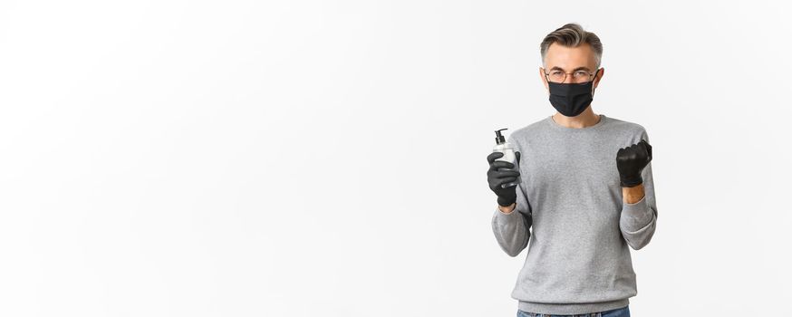 Concept of coronavirus, lifestyle and quarantine. Image of cheerful caucasian man in medical mask and gloves, showing hand sanitizer and making fist pump to rejoice, standing over white background.