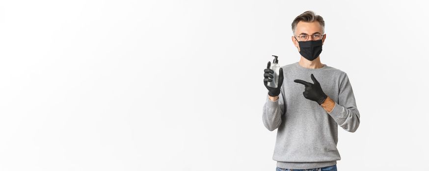 Concept of coronavirus, lifestyle and quarantine. Attractive middle-aged man in black medical mask and gloves, recommending use hand sanitizer, pointing at antiseptic, standing over white background.