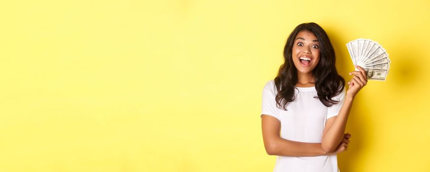 Portrait of beautiful african-american girl, smiling happy and showing money, going shopping, standing over yellow background.