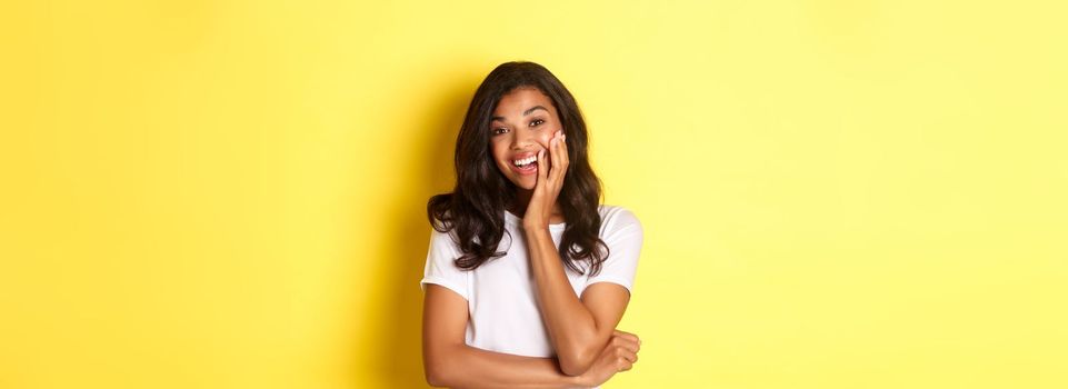Portrait of attractive african-american girl, looking amazed and smiling, standing over yellow background in casual white t-shirt.