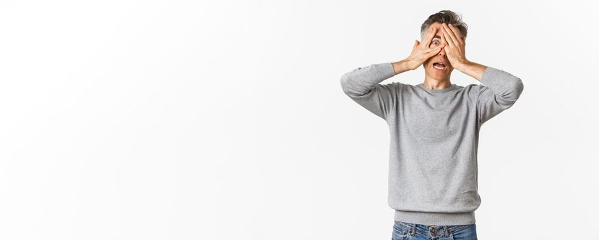 Image of embarrassed of scared middle-aged man cover his eyes with hands, peeking through fingers with shocked expression, standing over white background.