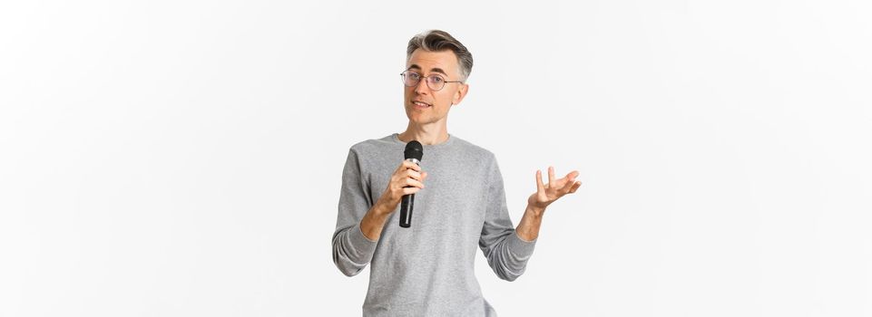 Portrait of handsome middle-aged man in glasses and gray sweater, making speech, talking with microphone and explaining something, standing over white background.