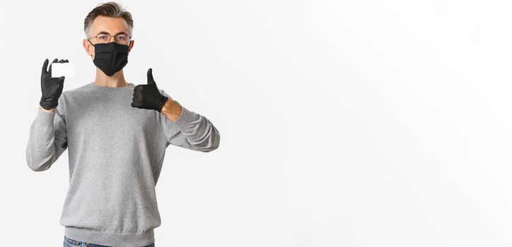 Concept of covid-19, social distancing and lifestyle. Satisfied middle-aged man in medical mask, gloves and glasses, recommending bank, showing credit card and thumbs-up.