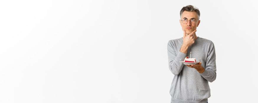 Image of thoughtful and serious middle-aged man, celebrating birthday, holding b-day cake and thinking what wish to make, standing over white background.