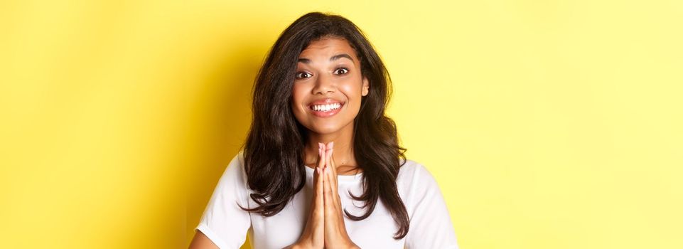 Close-up of cheerful african-american girl, smiling and saying thank you, press hands over chest in pray gesture, standing over yellow background.