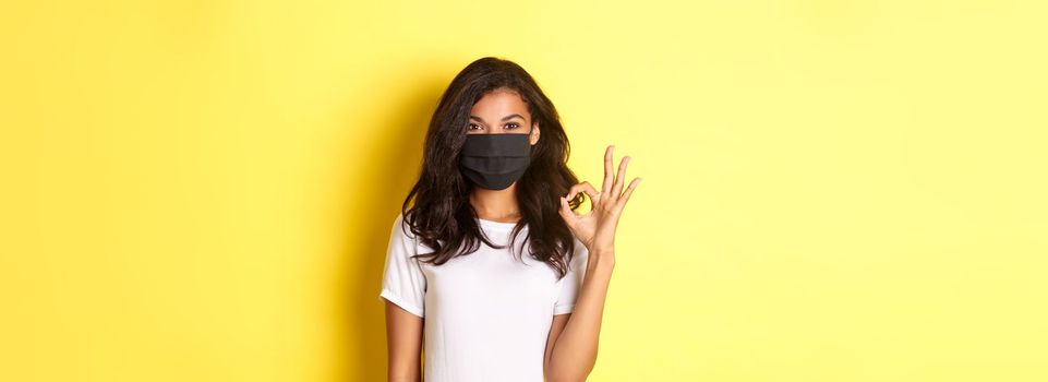 Concept of coronavirus, pandemic and lifestyle. Portrait of smiling african-american woman in face mask, showing okay sign in approval, recommend or guarantee something, yellow background.