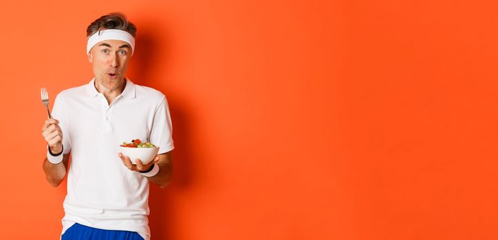 Concept of sport, fitness and lifestyle. Portrait of joyful middle-aged guy in workout uniform, holding fork and salad, eating healthy, standing over orange background.