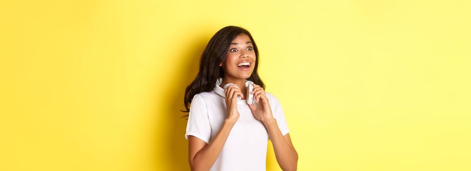 Portrait of beautiful african-american woman, holding headphones on neck and smiling, looking at upper left corner amazed, standing over yellow background.