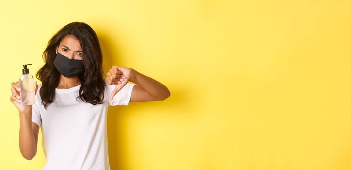 Concept of covid-19, social distancing and lifestyle. Image of disappointed african-american woman in face mask, showing thumbs-down, do not recommend bad hand sanitizer, yellow background.