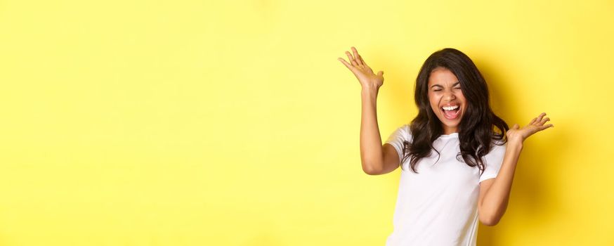 Portrait of cheerful african-american girl, shouting for joy and celebrating, achieve goal or winning something, standing over yellow background.