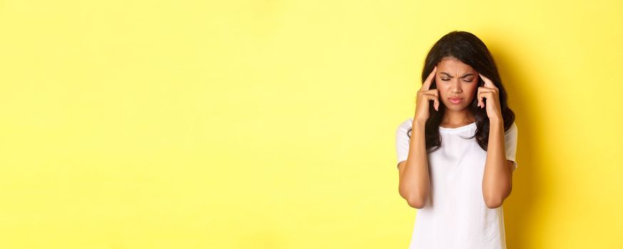 Portrait of young african-american female model feeling sick, frowning and touching head, complaining on headache, standing over yellow background.