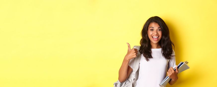 Image of cheerful african-american girl student, holding notebooks for classes and backpack, showing thumbs-up, like studying, standing over yellow background.