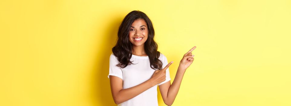 Portrait of cheerful and cute african american girl, pointing fingers left and smiling, showing advertisement on copy space, standing over yellow background.