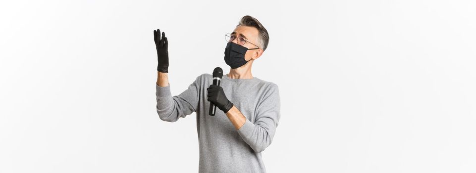 Concept of coronavirus, lifestyle and quarantine. Portrait of charismatic middle-aged man in medical mask and gloves, singing serenade in microphone, standing over white background.