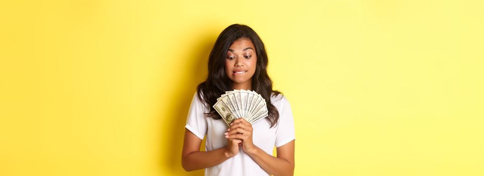 Portrait of attractive african-american woman, looking tempted at money, wanting to buy something, standing over yellow background.