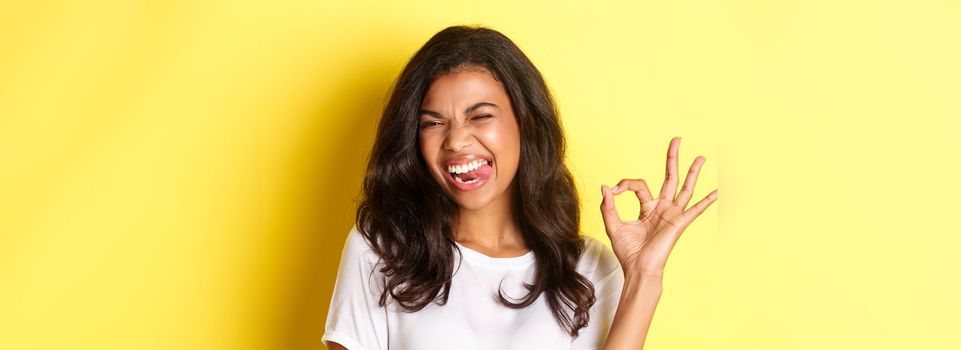 Close-up of cheerful african-american female model, showing okay sign, winking and looking satisfied, recommending something good, standing pleased over yellow background.