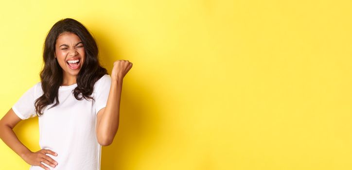 Portrait of happy african-american woman, triumphing and winning, making fist pump and shouting for joy, standing pleased against yellow background.