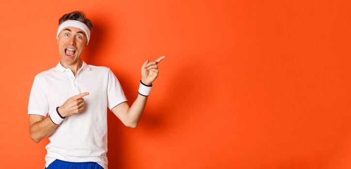 Concept of workout, sports and lifestyle. Image of excited middle-aged male athlete, wearing activewear for exercises, pointing fingers at upper right corner, showing logo, orange background.