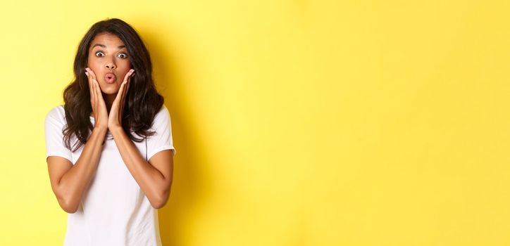 Portrait of attractive african-american teenage girl looking amazed, saying wow and stare at camera excited, standing over white background.