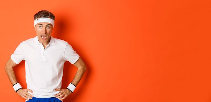 Concept of workout, sports and lifestyle. Portrait of startled middle-aged fitness guy in activewear, drop jaw and looking at camera amazed, standing over orange background.