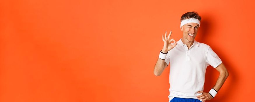 Concept of sport, fitness and lifestyle. Portrait of confident middle-aged sportsman, showing okay sign and look pleased, guarantee something or recommending gym, orange background.