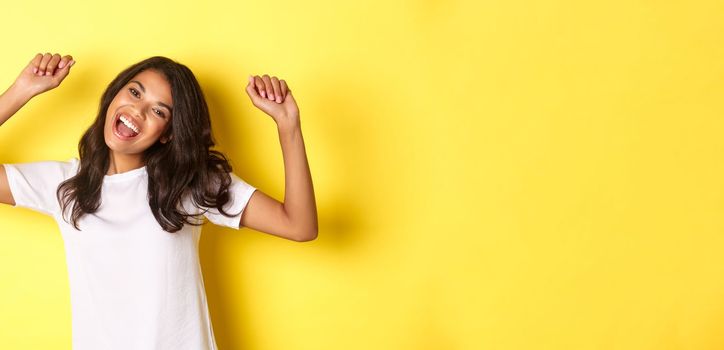 Image of cheerful african-american girl winning, looking happy and celebrating victory, triumphing about achievement, standing over yellow background.