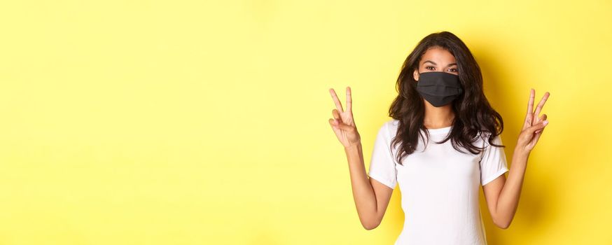 Concept of covid-19, social distancing and lifestyle. Portrait of beautiful african-american girl in black face mask, showing peace signs and smiling, standing over yellow background.