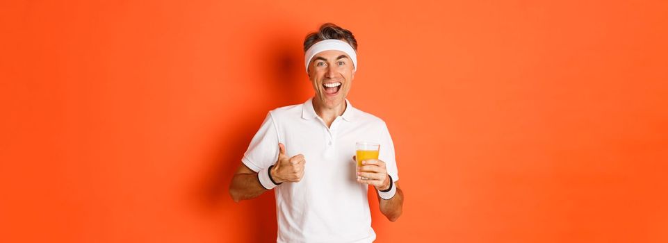 Concept of workout, gym and lifestyle. Image of active and healthy middle-aged sport guy, showing thumbs-up and drinking juice, smiling happy, standing over orange background.