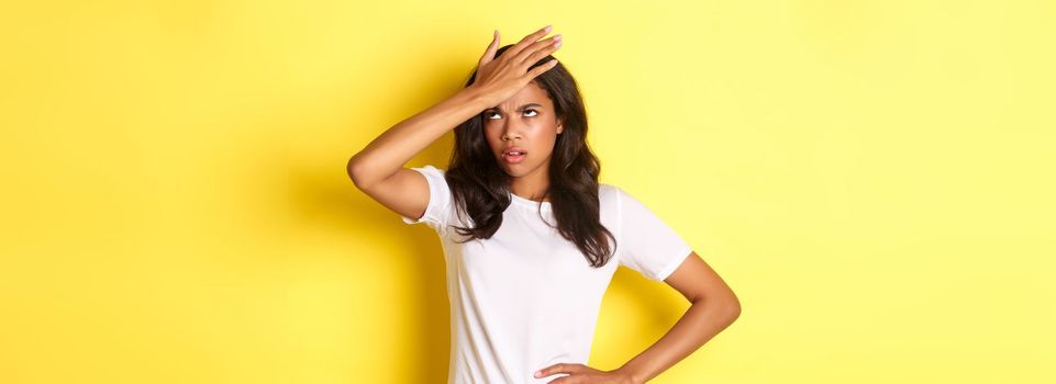 Portrait of annoyed african-american girl, slap forehead and roll her eyes bothered, forgot something important, standing over yellow background.