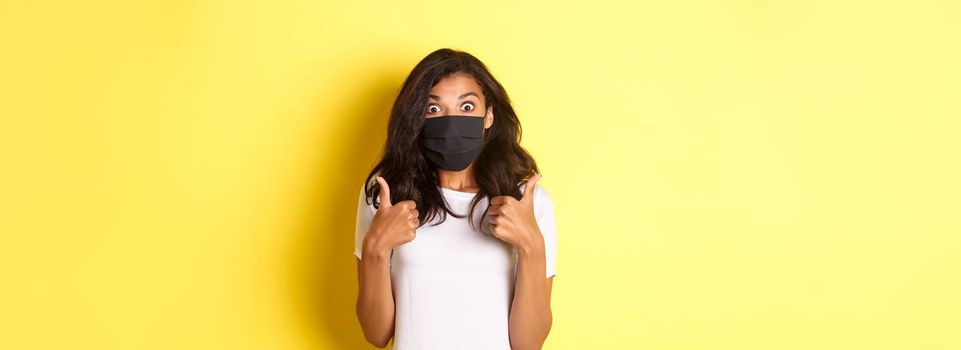 Concept of coronavirus, pandemic and lifestyle. Portrait of beautiful african-american girl in black face mask, showing thumbs-up and looking amazed, recommending something, yellow background.