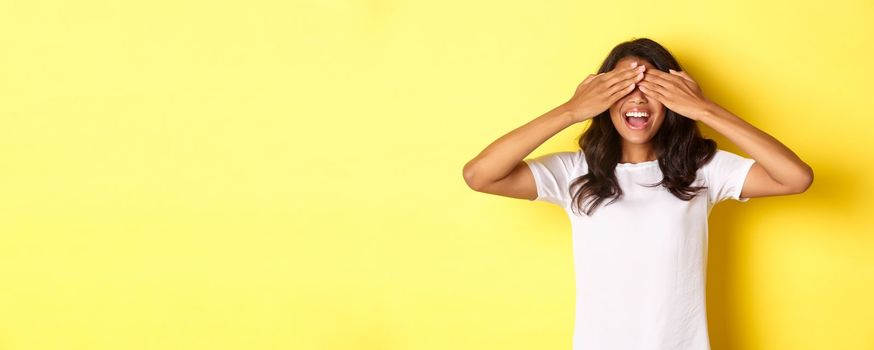 Image of excited african-american girl waiting for surprise, smiling and covering eyes with hands, standing over yellow background.