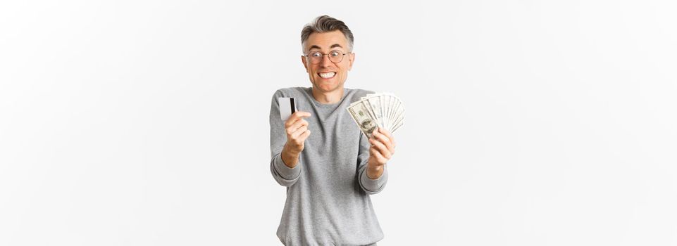 Portrait of excited and happy middle-aged man, looking amazed at credit card and money, standing over white background.