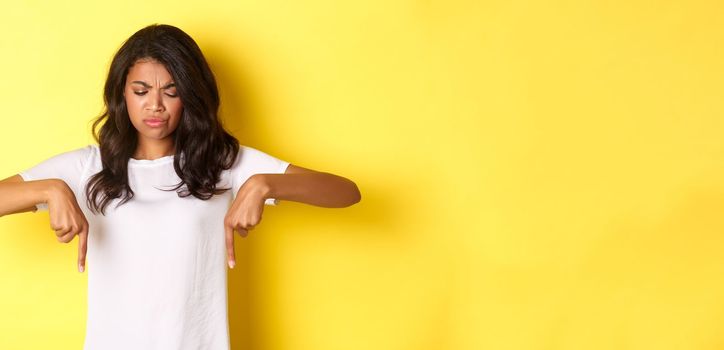 Portrait of sad and disappointed african-american girl, complaining about something bad, looking and pointing fingers down with displeased face, standing over yellow background.