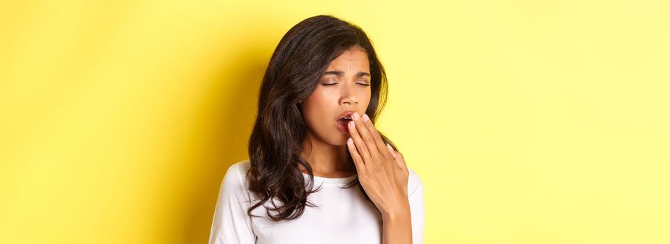 Close-up of tired and sleepy african-american girl, yawning and looking exhausted, standing in white t-shirt over yellow background.