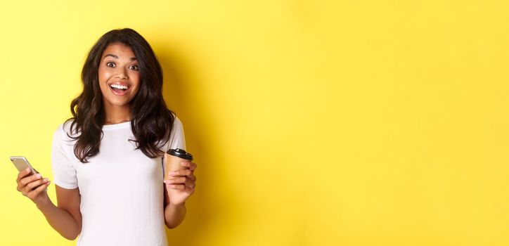 Portrait of attractive african-american girl smiling, holding coffee cup and smartphone, standing over yellow background.