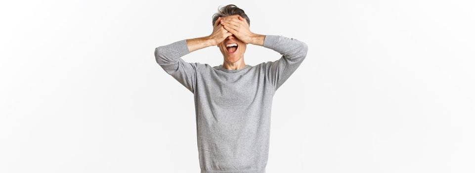 Portrait of happy middle-aged man waiting for surprise, shut eyes and smiling, playing hide n seek, standing over white background.