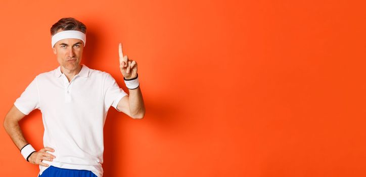 Portrait of serious middle-aged male athlete, wearing sportswear, pointing finger up and showing logo about sports and gym, standing over orange background.