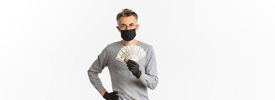 Concept of covid-19, social distancing and lifestyle. Portrait of successful middle-aged man in medical mask, gloves and glasses, showing money, standing over white background confident.