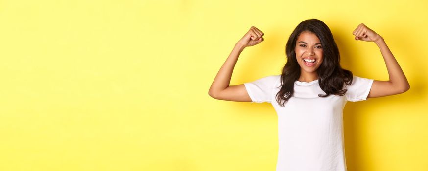 Image of confident and cheerful african-american girl, flexing biceps and smiling, looking strong and assertive, standing over yellow background.